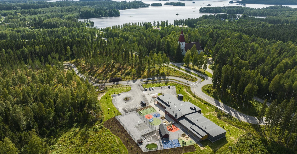 An aerial view of a kindergarten surrounded by a forest. The church and the lake can be seen in the background.