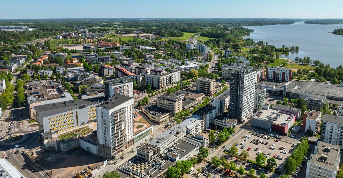 Ilmakuva kaupungista, jossa kerrostalo ja toimitilarakennuksia, taustalla vesistö, ympärillä metsikköä